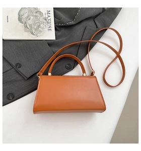 Flat lay of a brown trapezoid leather handbag styled with a grey blazer and accessories