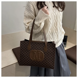 Close-up of brown tote bag with M-letter print and leather straps on white sweater