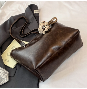 Flat lay of brown tote bag with plush cat charm on grey blazer and white fabric background