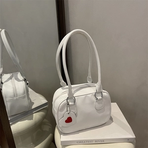 Sac Boston blanc à motif litchi avec pendentif cœur rouge posé sur un livre