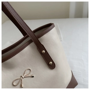 Close-up of beige leather tote bag featuring dark brown trim and gold bow metal detail