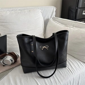 Large black matte leather tote bag with gold bow accent on a white sofa setting
