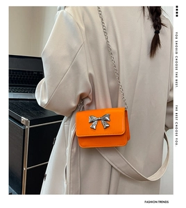 Close-up of orange leather mini bag with silver metal bow and chain strap on beige coat