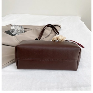 Détail d'un grand sac cabas marron foncé avec charm ours en peluche sur un lit blanc
