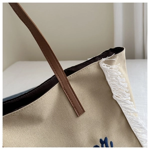 Détail du sac cabas en toile montrant la poignée en cuir brun et les franges en coton