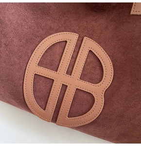 Detail shot of a dark reddish-brown suede tote bag with a stitched leather logo patch