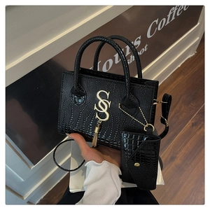 Detalle de bolso negro con diseño de cocodrilo, logo S dorado y borla en un café