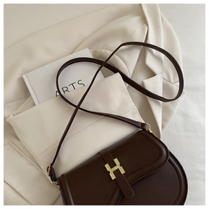 Detail of dark brown leather saddle bag featuring a gold H-shaped buckle on a beige blazer