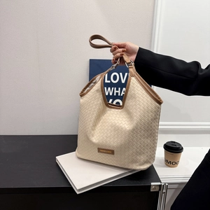 Large beige woven tote bag with brown leather handles and brand label on a desk