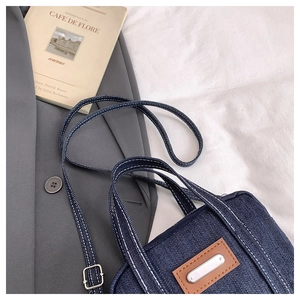 Detail of blue denim pillow bag showing white stitching and adjustable strap on a grey blazer