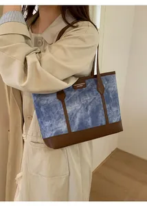 Detail of blue denim tote bag with brown leather handles worn by a model in a trench coat