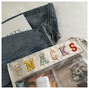 Close-up of clear PVC cosmetic bag with colorful SNACKS embroidery and white leather trim