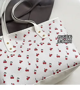 Close-up of white tote bag with red cherry prints, litchi texture and gold rivet detail