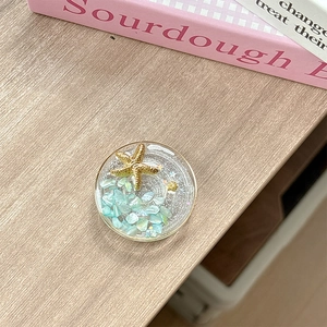 Detail of round ocean-themed phone grip with gold stars and blue stones on wooden surface