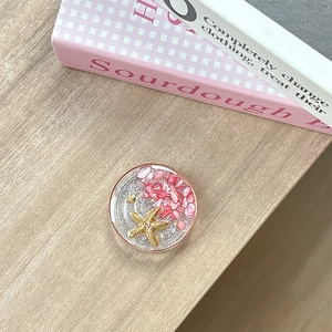 Detail of round phone grip featuring pink crushed stones and golden starfish seashell elements