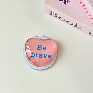 Detail einer rosa Herz-Handyhalterung mit 'Be brave' Aufdruck auf hellem Schreibtisch