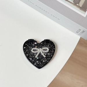 Close-up of black glitter heart-shaped compact mirror with a rhinestone bow on white desk