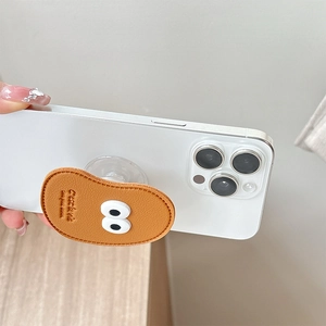 Brown leather phone grip with cartoon eyes held on white smartphone against a gray wall