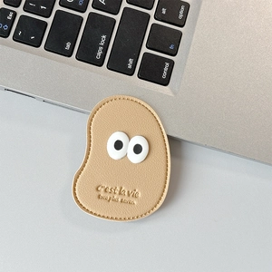 Close-up of beige leather bean-shaped phone grip with large eyes and C'est la vie text