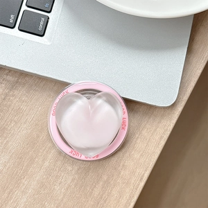Detail of a pink heart-shaped phone grip with Good Luck text on a wooden desk near a laptop