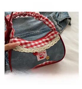 Close-up of denim shoulder bag featuring red plaid trim, lace detail, and cherry patch