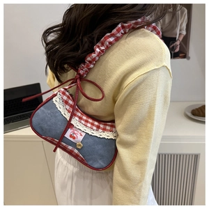 Detail of denim underarm bag featuring red plaid straps and lace trim with cherry patch