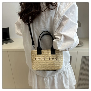 Detail view of woven straw tote bag with black shoulder strap and embroidered lettering