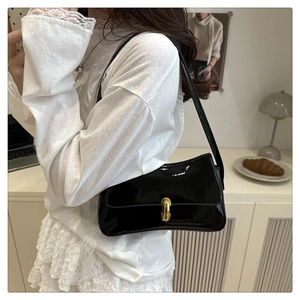 Glossy black patent leather underarm bag with gold turn-lock worn by a model in a minimal room