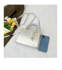 Silver square crossbody bag with pearl bow charm next to mobile phone for size comparison