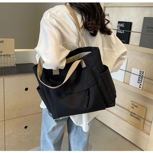 Detail of a large black canvas tote bag worn by a model in a minimalist wooden room