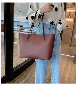Deep burgundy tote bag with chain strap shown on a model in a modern interior setting