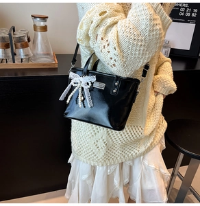 Detail of black glossy bucket bag with white lace bow, pearl pendant and metal plaque