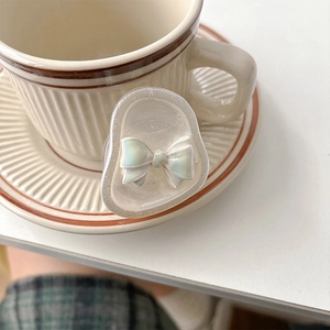 Pearly iridescent bow cell phone grip stand with translucent resin finish on a coffee saucer