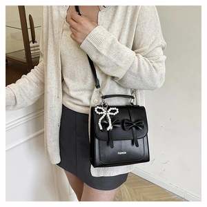 Detail of a black leather crossbody bag featuring bows and a white pearl charm