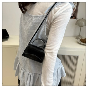 Minimalist black glossy leather square crossbody bag shown worn by a model in a modern home