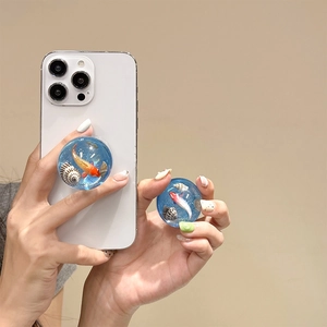 Macro detail of a 3D goldfish and seashell resin phone grip on a white smartphone
