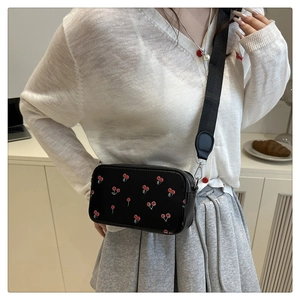 Detail of black crossbody bag with red cherry print and wide nylon strap featuring metal buckles