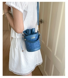 Detail of blue denim bucket bag with gold lettering shown on a white lace dress