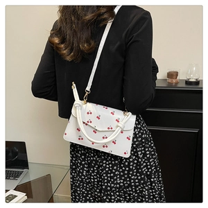 Detail view of a white crossbody bag featuring red cherry prints and knotted top handle
