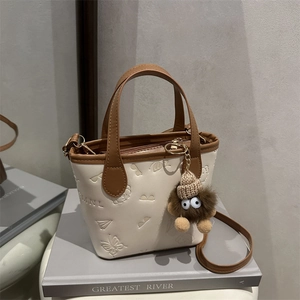 Beige bucket bag with brown handles, embossed patterns and a plush charm on books