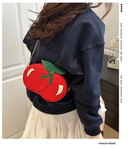 Detail of red cherry-shaped crossbody bag with green leaf and gold chain on model