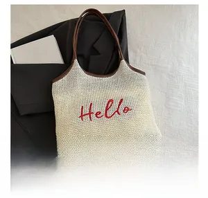 Detail of woven straw tote bag featuring red Hello embroidery and brown leather handles