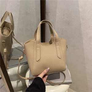 Minimalist beige pebble leather handbag with dual handles and crossbody strap on pedestal