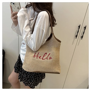 Detail of woven straw tote bag featuring red Hello embroidery and brown leather shoulder straps