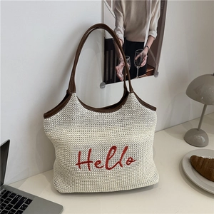 Beige woven straw tote bag with red Hello embroidery and brown leather straps on a desk
