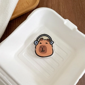 Close-up of a cute cartoon capybara phone grip with blue headphones in a white tray