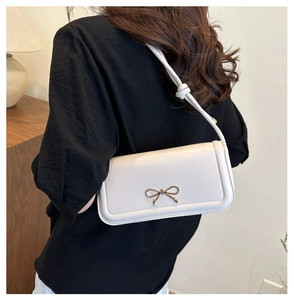 Detail of white underarm bag featuring a golden metal bow and adjustable knotted strap