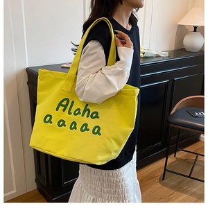 Detail of yellow canvas tote bag with green text, worn by model in cozy interior setting