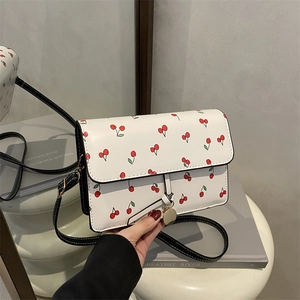White crossbody bag with red cherry print, black strap and gold pendant in a minimalist room