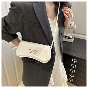 Close-up of cream crossbody baguette bag featuring a gold metal bow and smooth texture